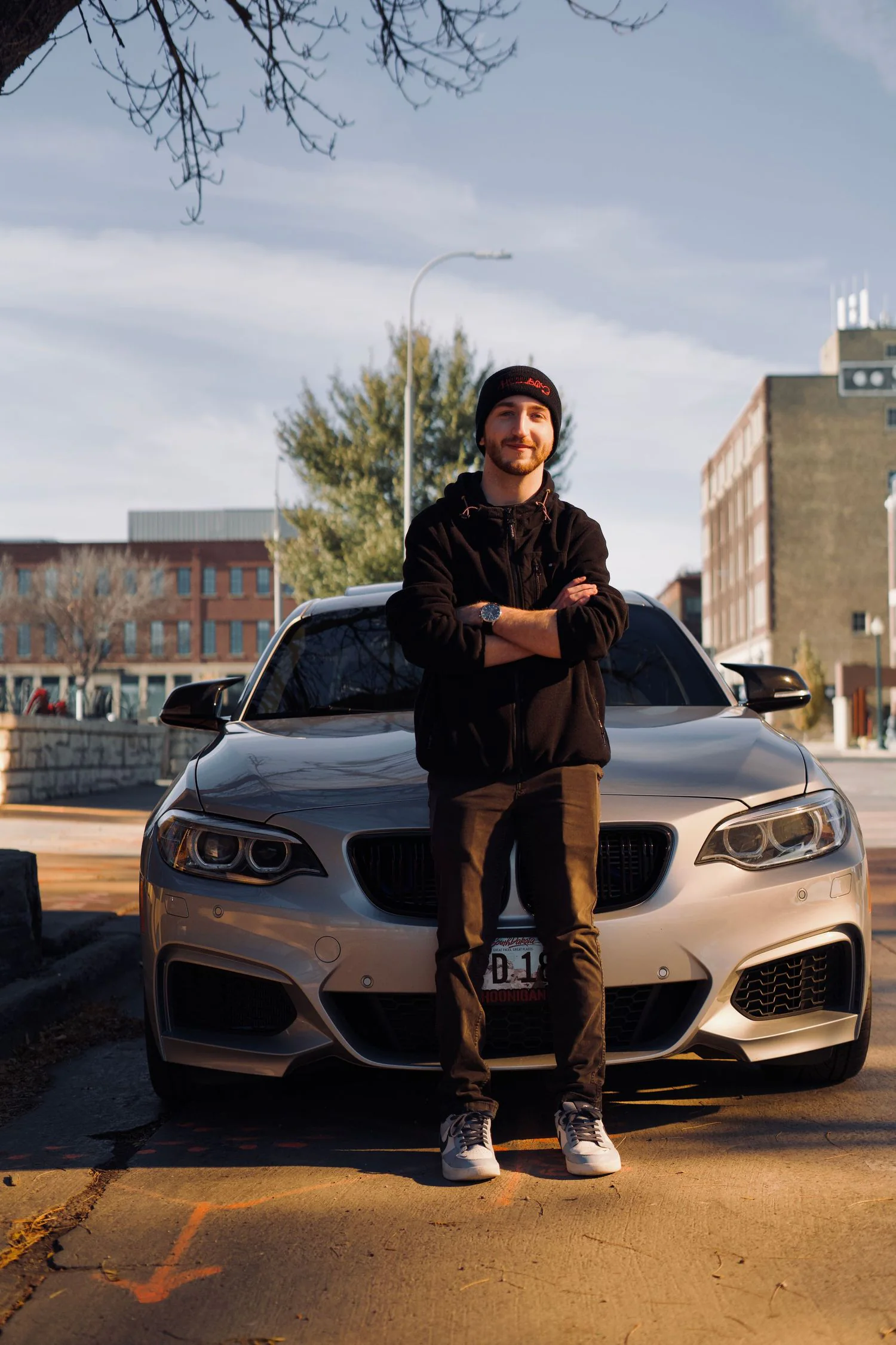 A man in black standing in front of a silver sports sedan in an urban parking area.