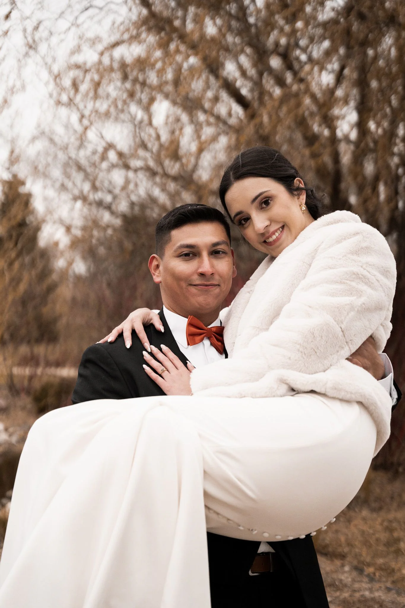 A groom carrying a smiling bride outdoors beneath bare trees.