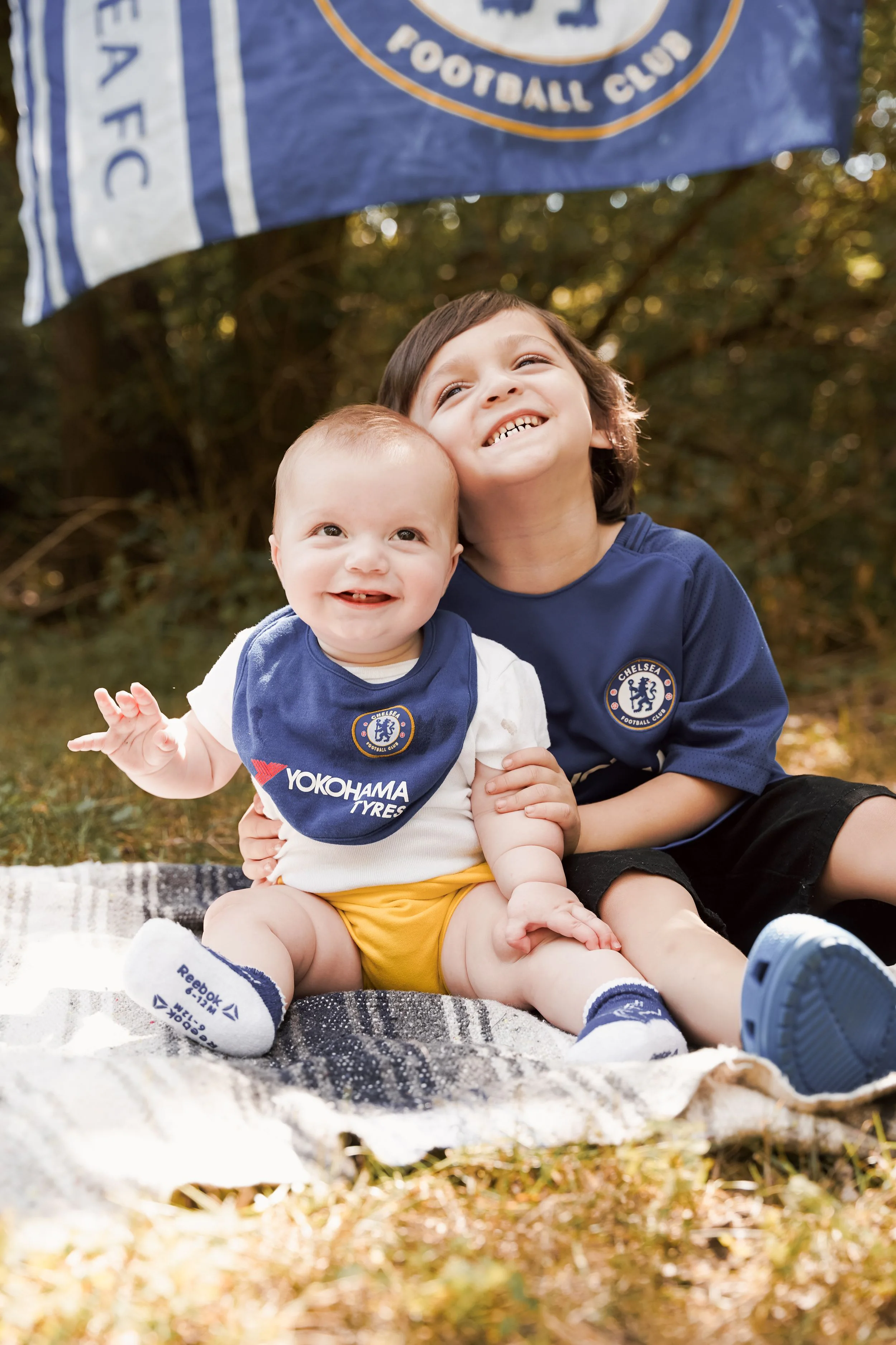 A woman holding a smiling baby outdoors near a sports-field banner.